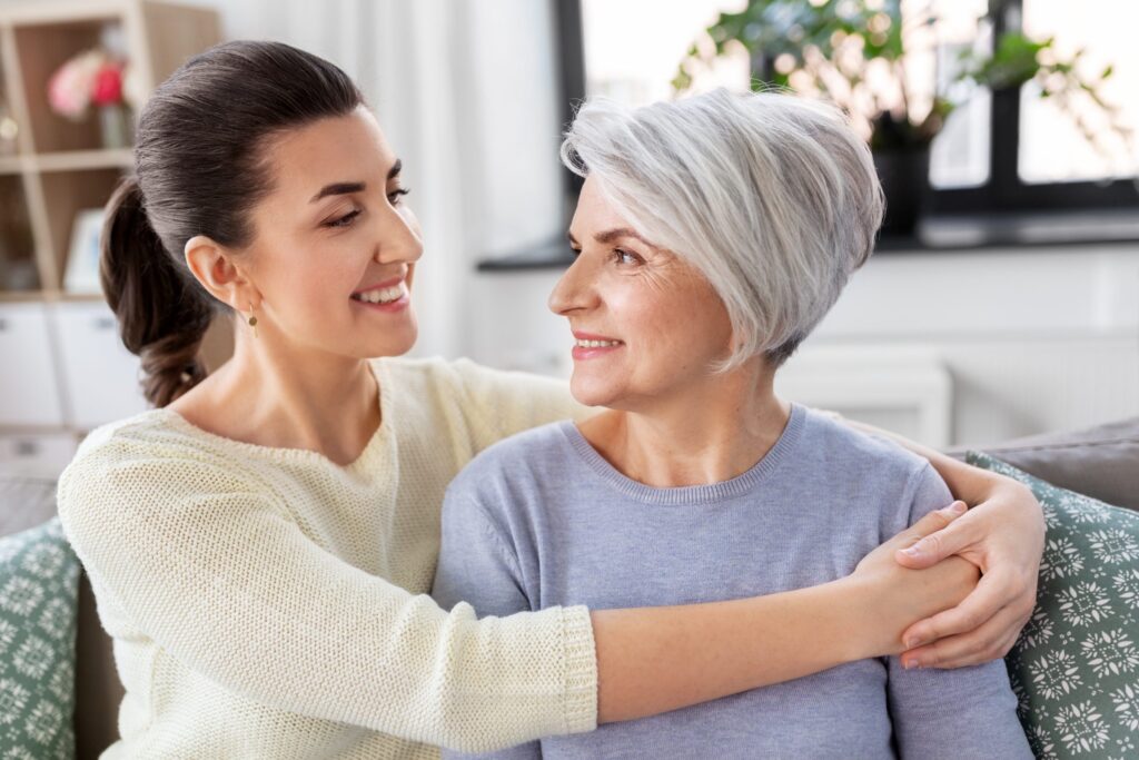In a well-lit room adorned with plants, a woman with gray hair and a smile is seated on the couch, embraced by a younger dark-haired woman. Their warm connection embodies the care found through Brookfield Elder Law.