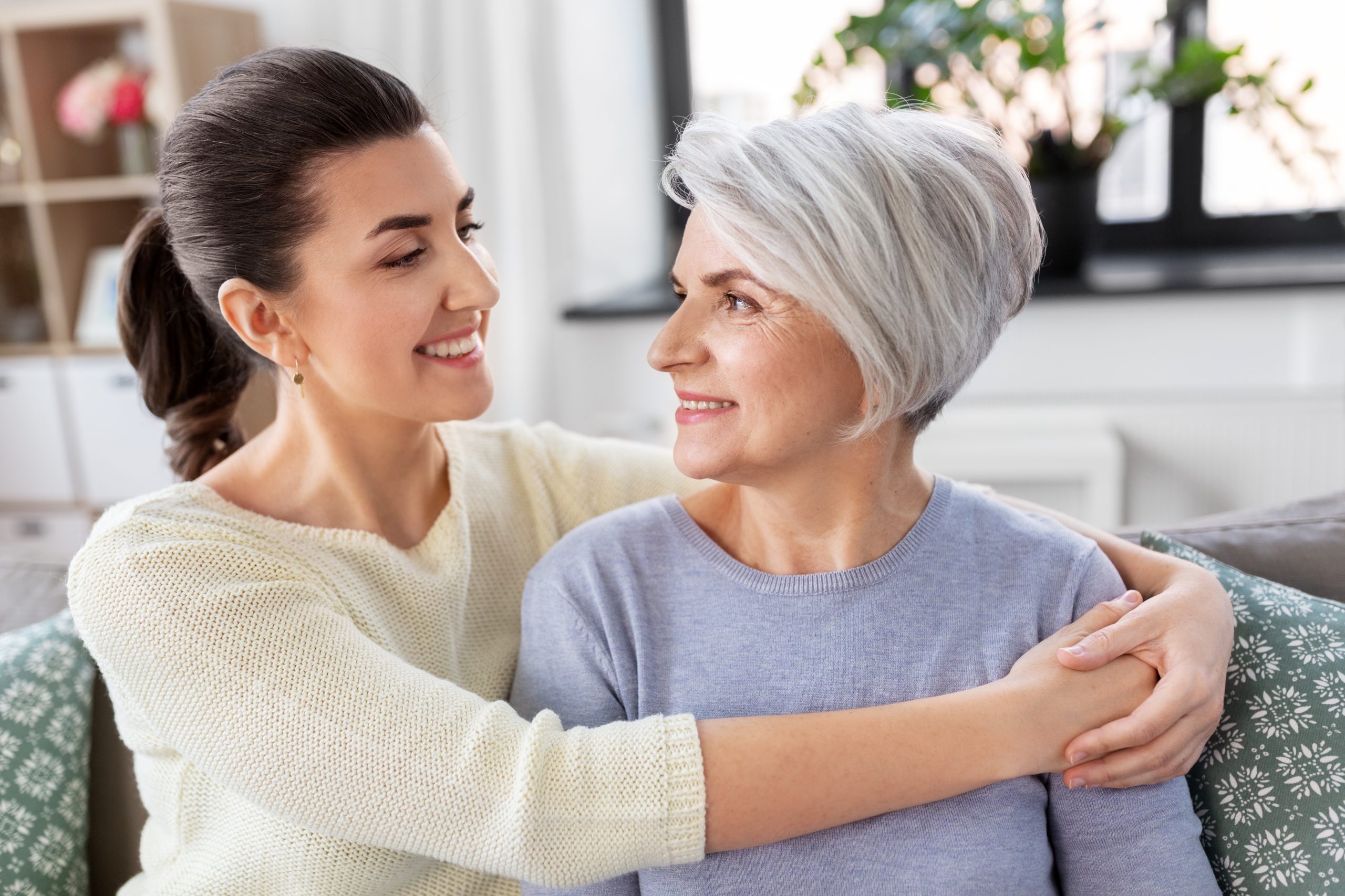 In a well-lit room adorned with plants, a woman with gray hair and a smile is seated on the couch, embraced by a younger dark-haired woman. Their warm connection embodies the care found through Brookfield Elder Law.
