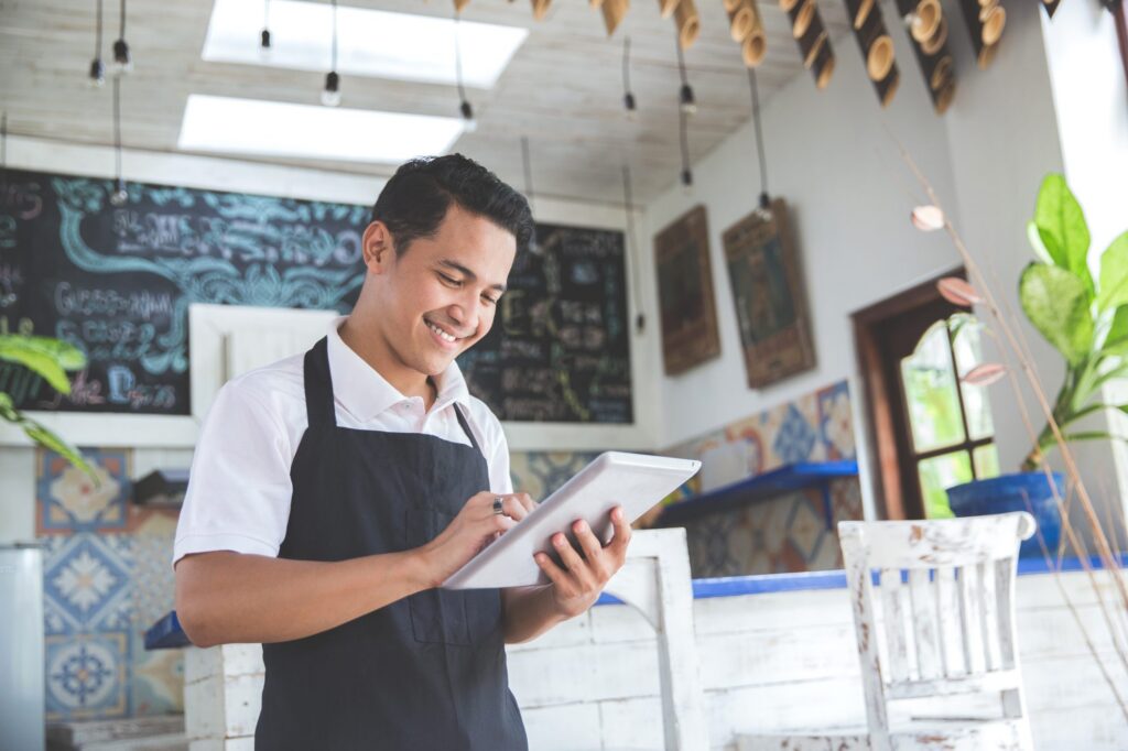 A young man wearing an apron smiles while writing on a notepad inside a bright, rustic café as he's happy that he planned his small business estate with a Deerfield attorney.