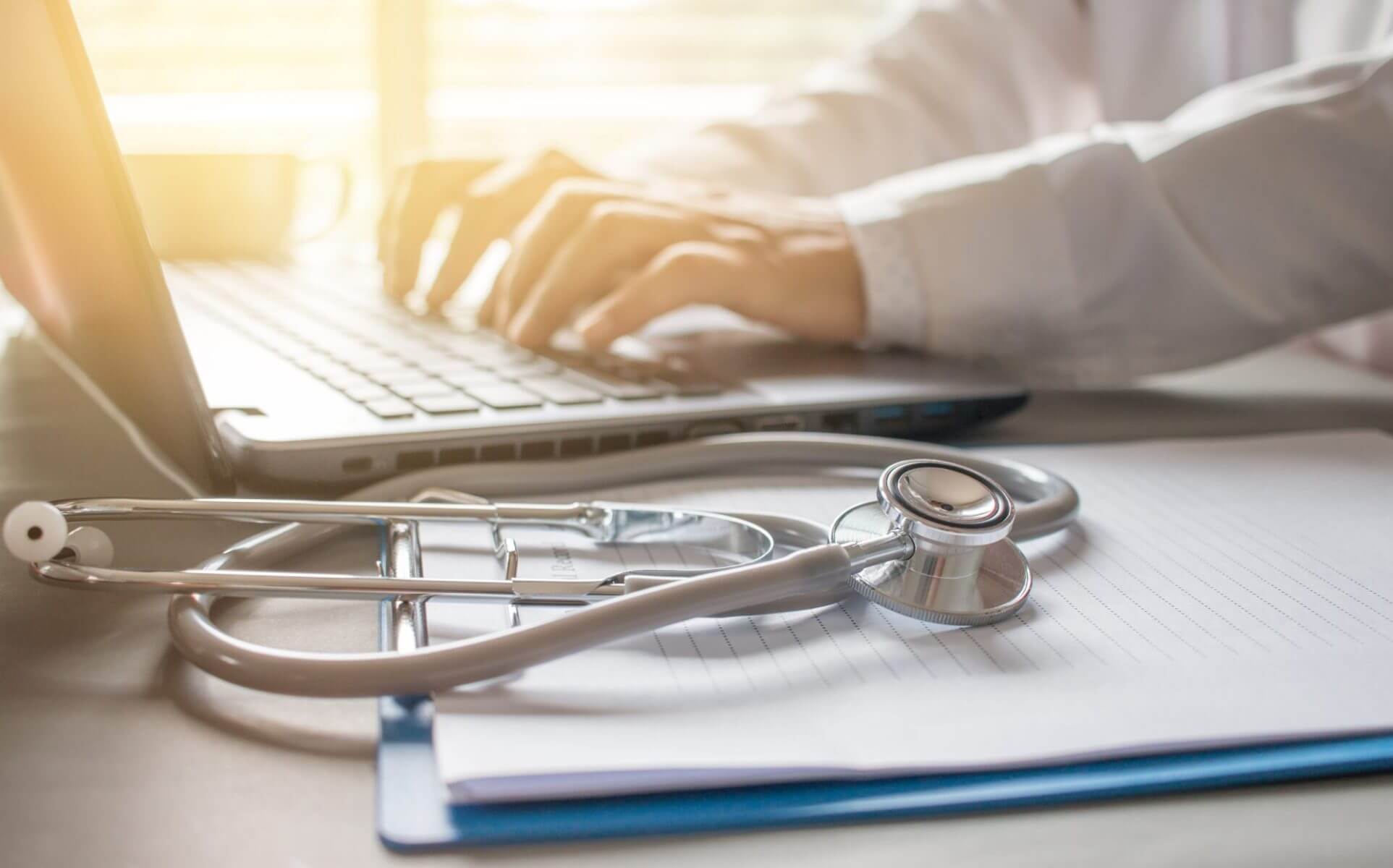 A stethoscope rests on a notebook beside a clipboard, echoing the precision of LaGrange Park Trusts. In the background, a person types on a laptop, crafting meticulous plans akin to Western Springs Estate Administration.