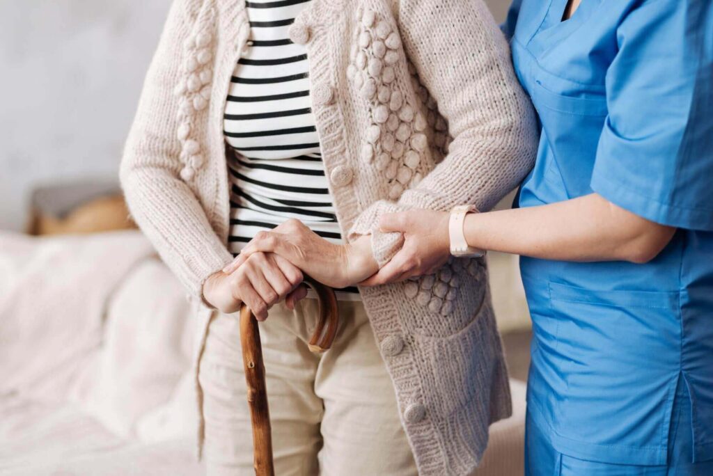 A caregiver in blue scrubs from Brookfield Elder Law assists a person with a cane, who is wearing a knitted cardigan and striped shirt.