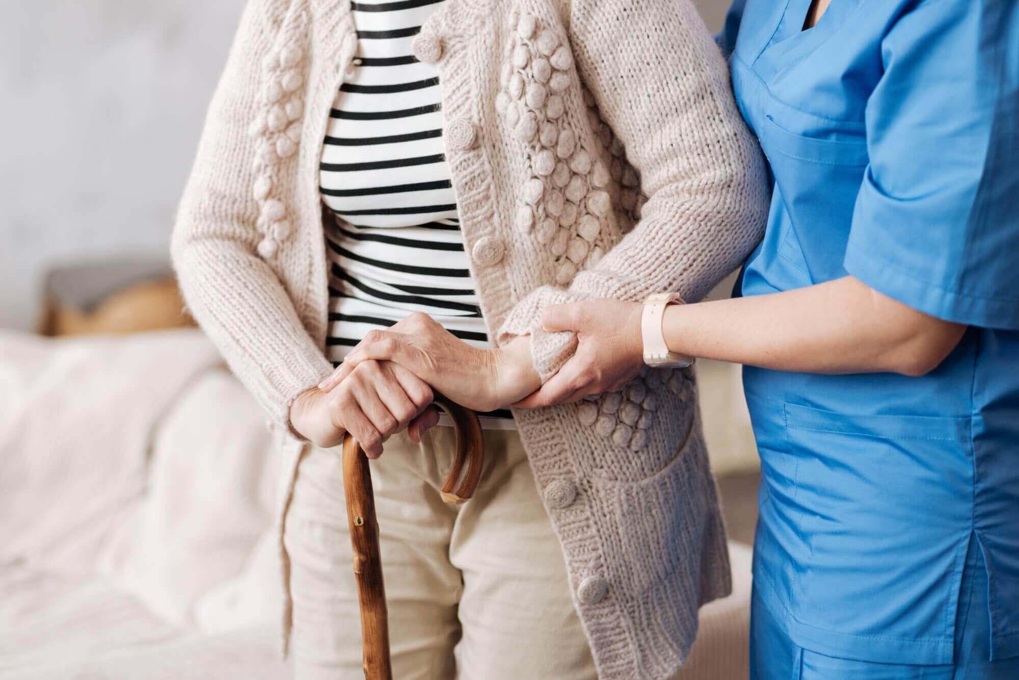 A caregiver in blue scrubs from Brookfield Elder Law assists a person with a cane, who is wearing a knitted cardigan and striped shirt.