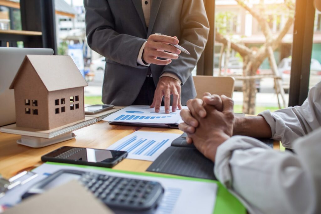 In a meeting room, two individuals discuss charts on the table. A small model house, calculator, and smartphones are visible. One person gestures with a pen, while discussing Western Springs Estate Administration, as the other listens intently with hands clasped.