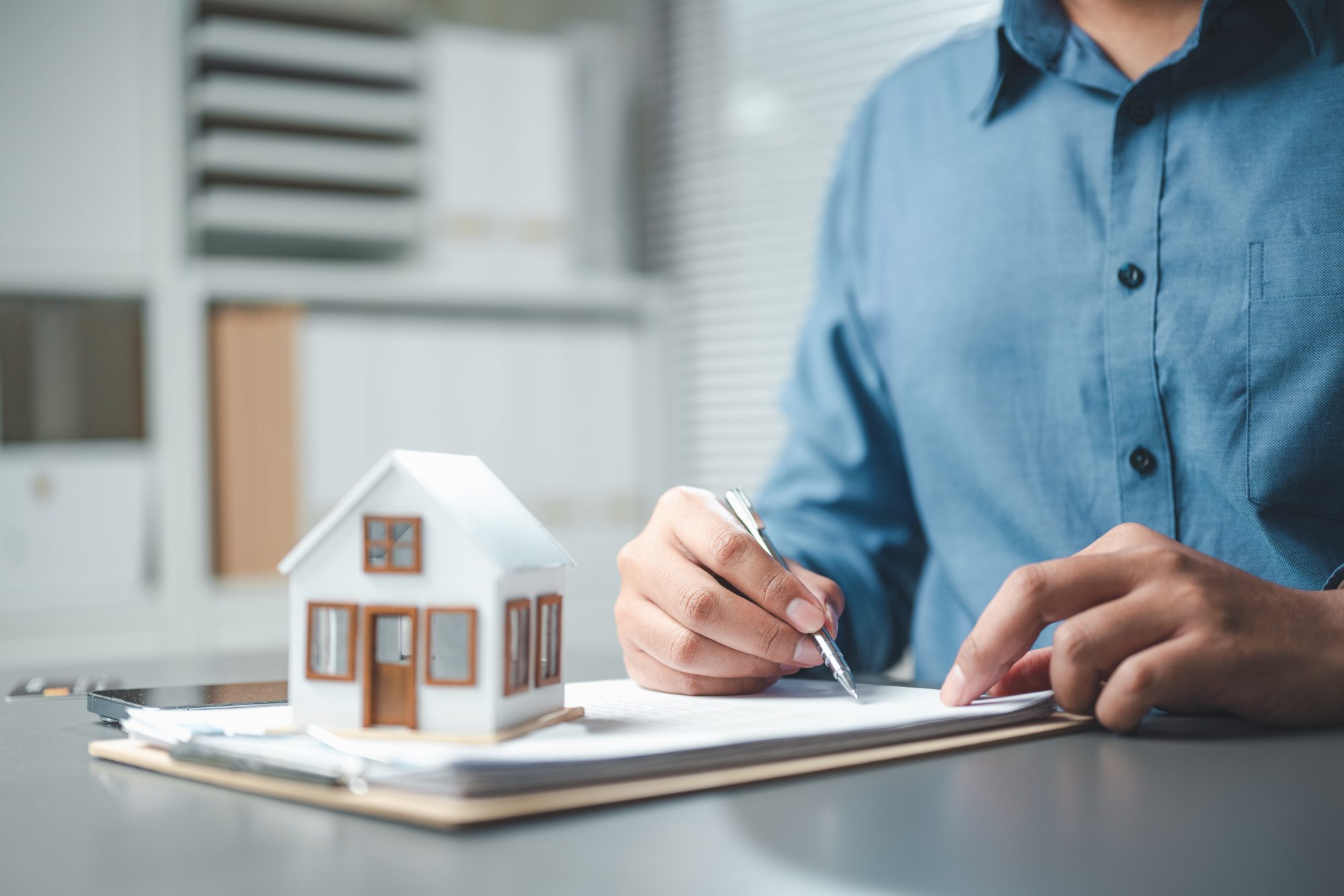 Person in a blue shirt signing documents at a desk with a small model house and papers, representing real estate or home ownership.