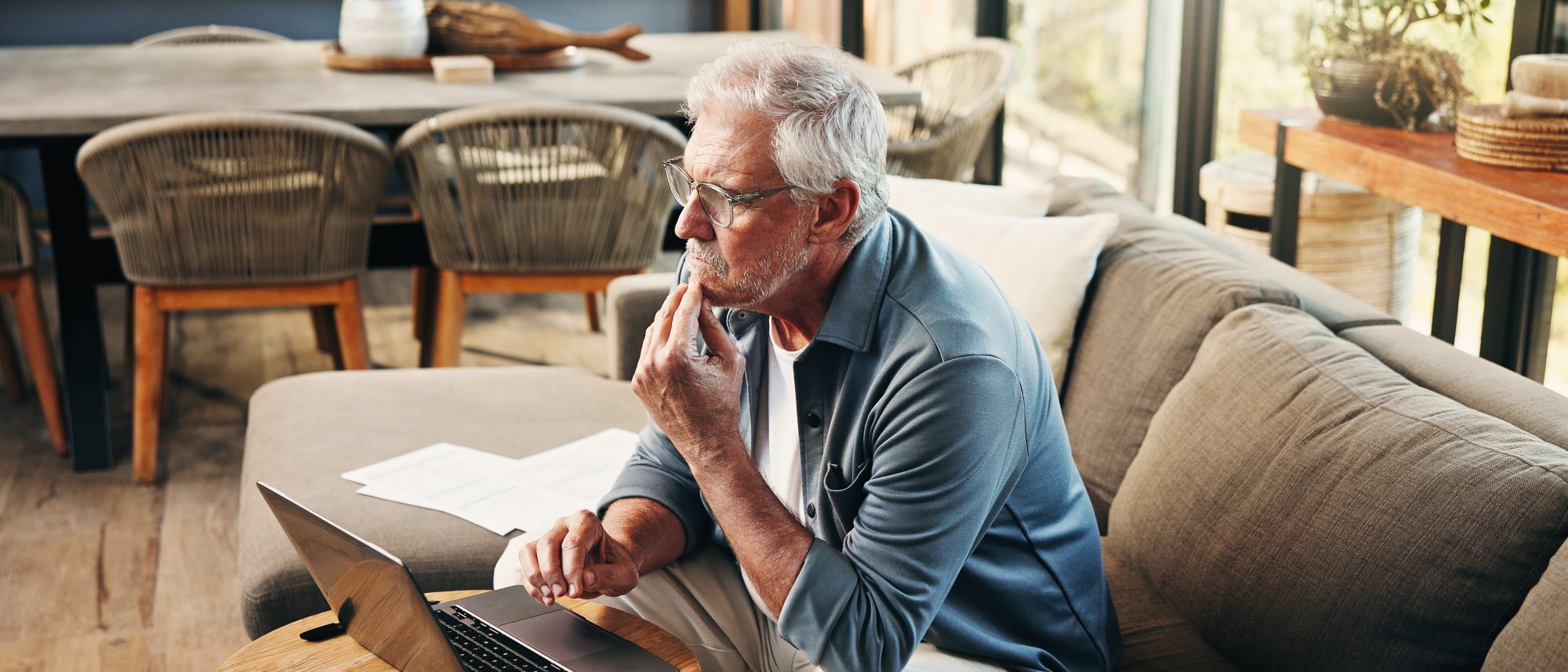 An older man sits on a sofa using a laptop, appearing thoughtful as he reviews papers related to his future with guidance from a La Grange estate planning lawyer in a well-lit living room.