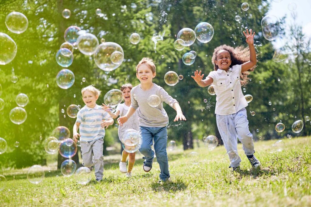 Four children joyfully run through a grassy field at the historic Hinsdale Wills estate, surrounded by bubbles on a sunny day.
