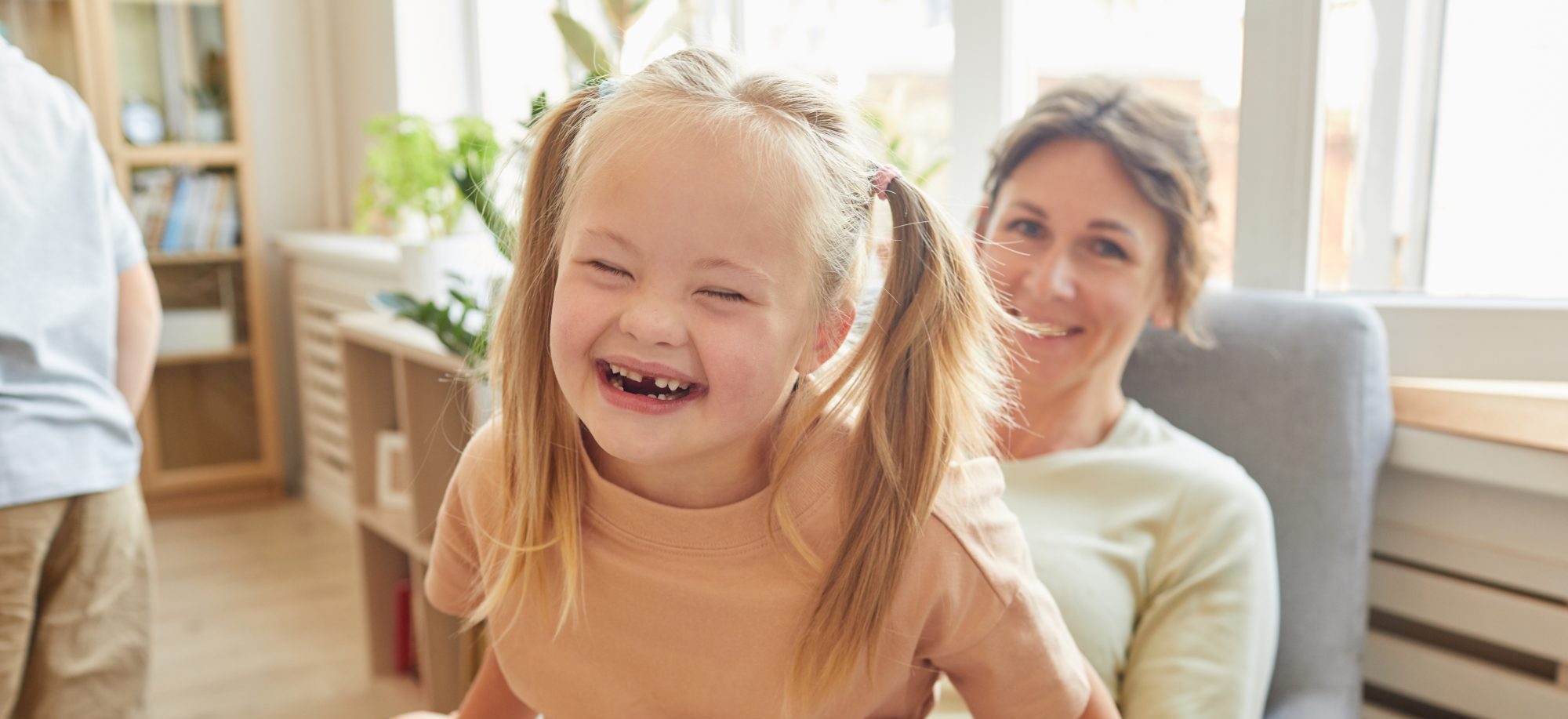 A smiling young girl with Down syndrome sits on a woman's lap in a bright room, holding a book, both looking toward the camera.