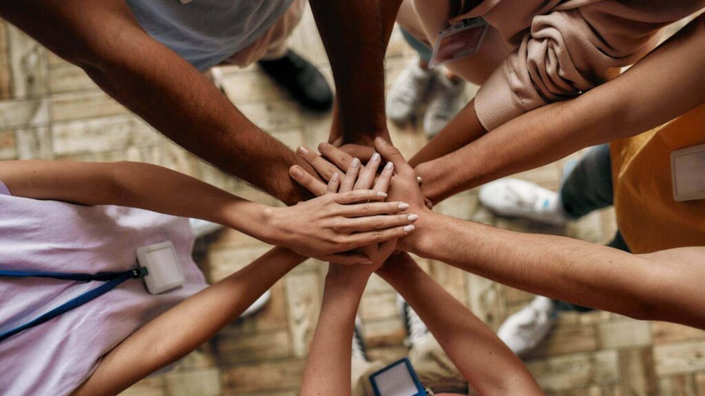 A diverse group of people stands in a circle on the wooden floor of the Western Springs Estate Administration office, each extending a hand to the center, overlapping their hands in a gesture of unity.