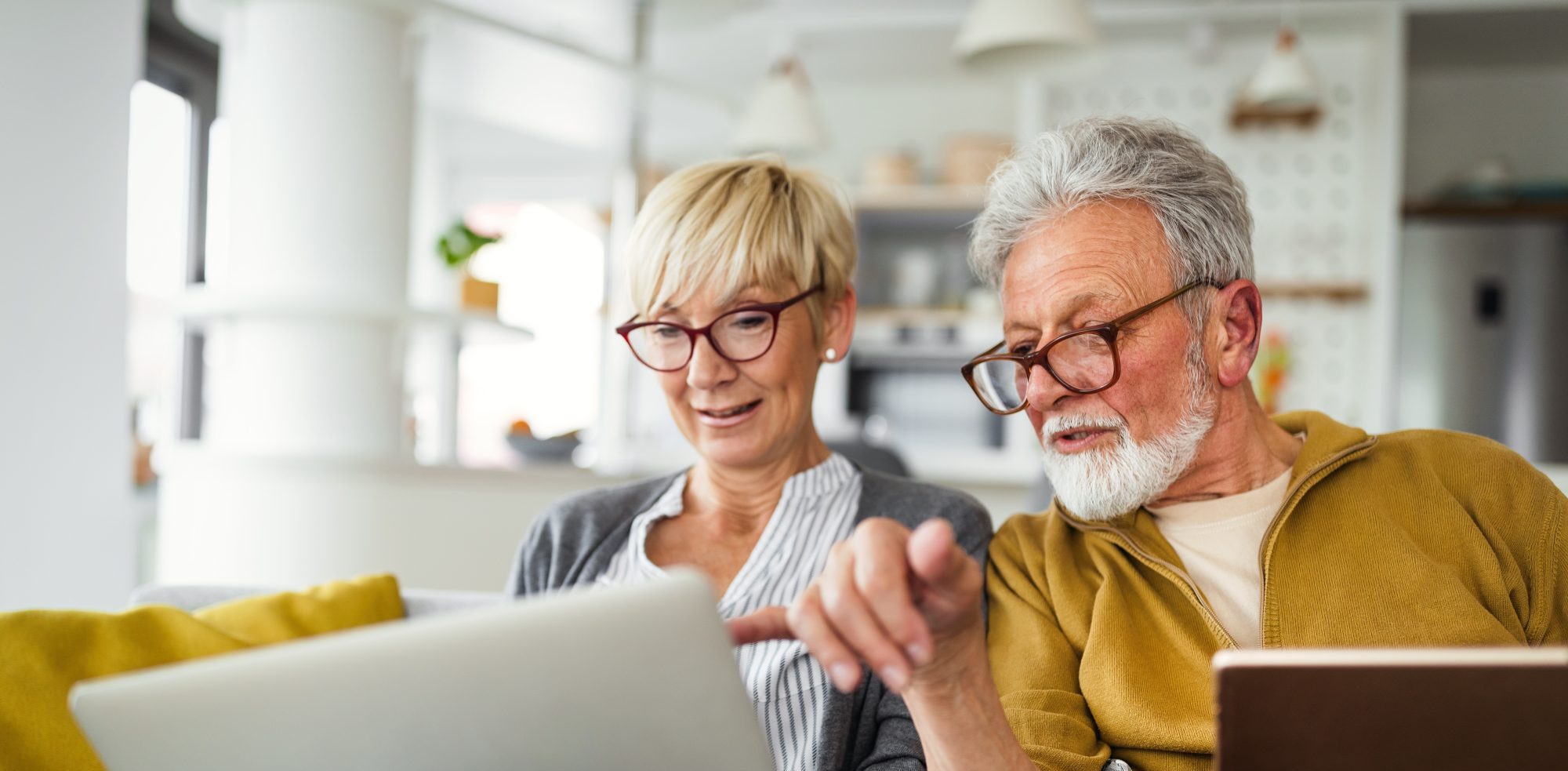 Two older adults sit on a couch, looking at a laptop together. The woman holds the laptop while the man, holding a tablet, points at the screen—perhaps researching Willow Springs elder law attorneys for trusted advice.