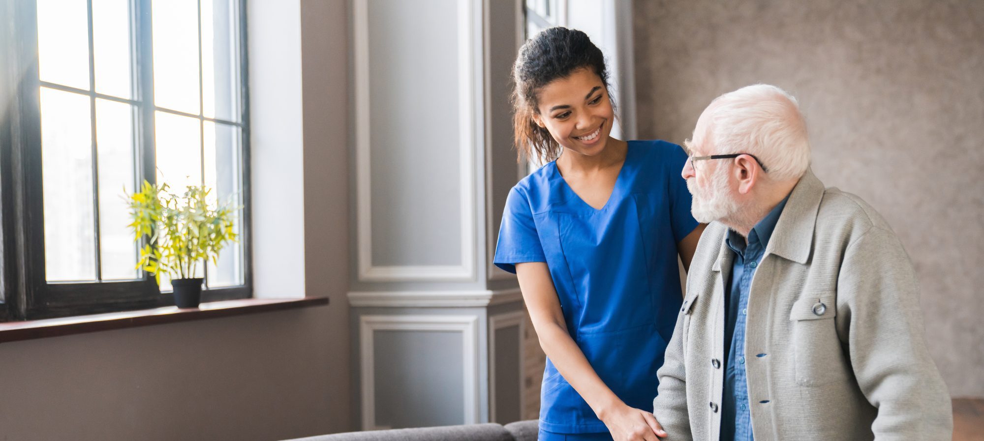 A caregiver in blue scrubs assists an elderly man with a walker beside a large window at Westchester Probate, ensuring every step reflects trust and care.