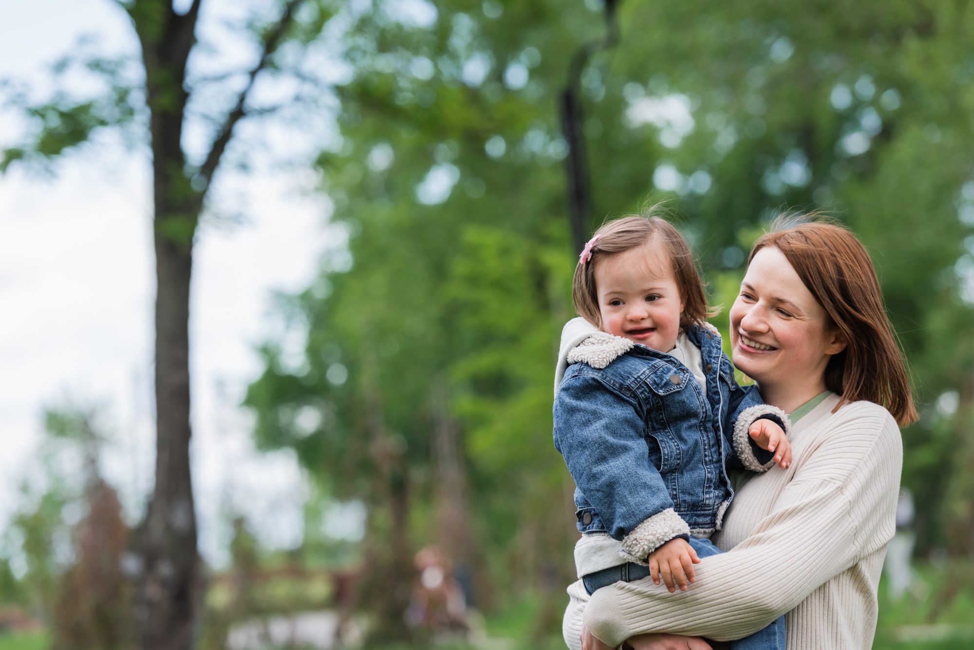 A woman smiles while holding a young girl wearing a denim jacket outdoors in a park with green trees in the background.