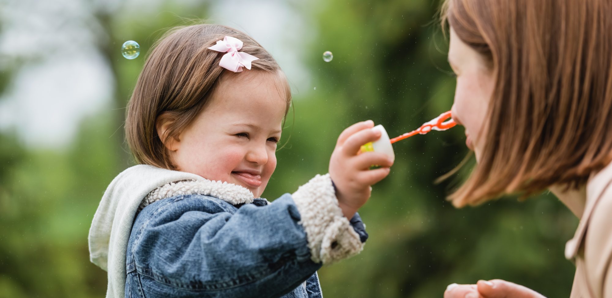 A young girl with Down syndrome smiles as she holds a bubble wand toward an adult woman outdoors, with blurred green trees in the background.
