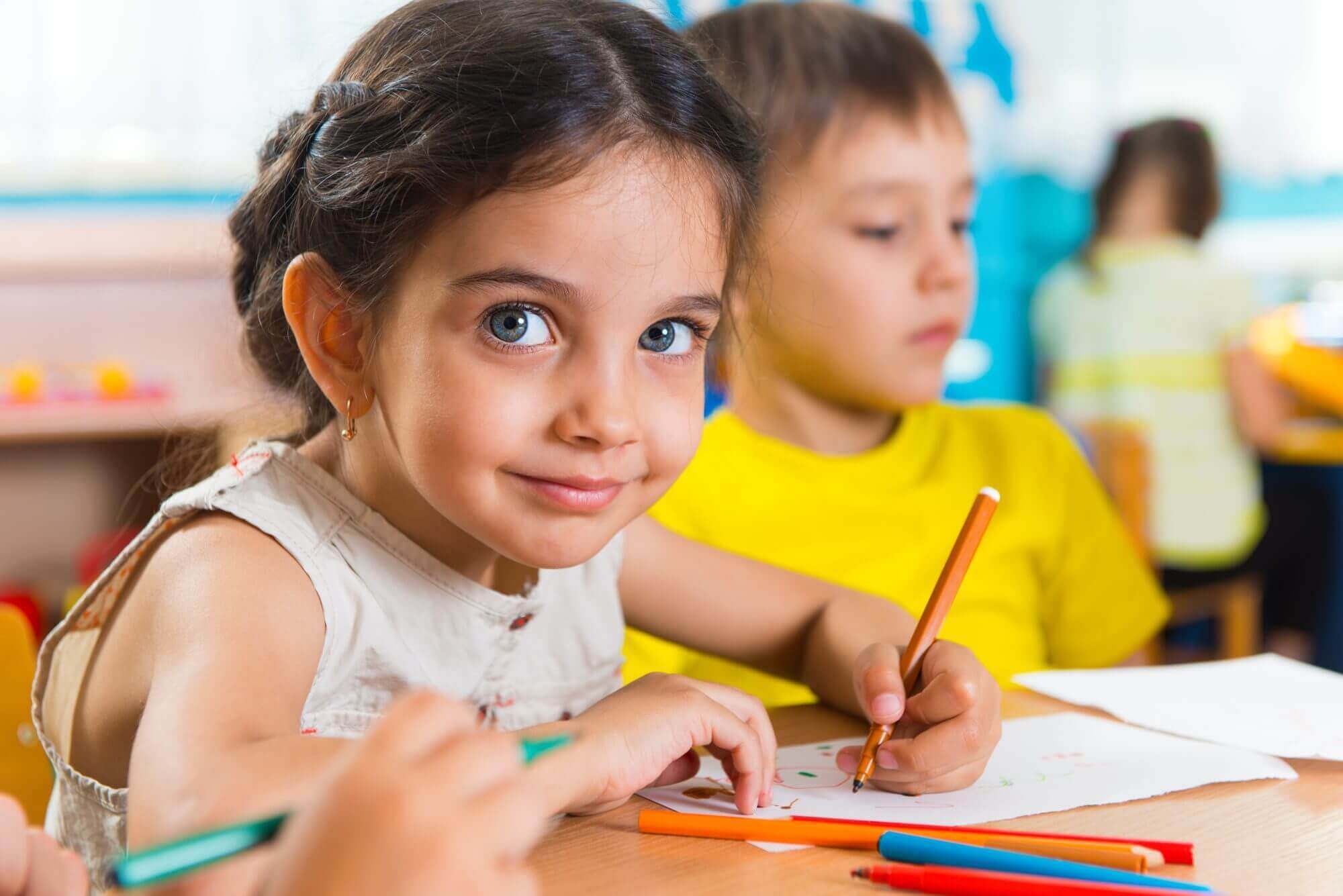 A young girl with brown hair draws with colored pencils at a table in a Western Springs estate administration office, where other children are visible in the background.