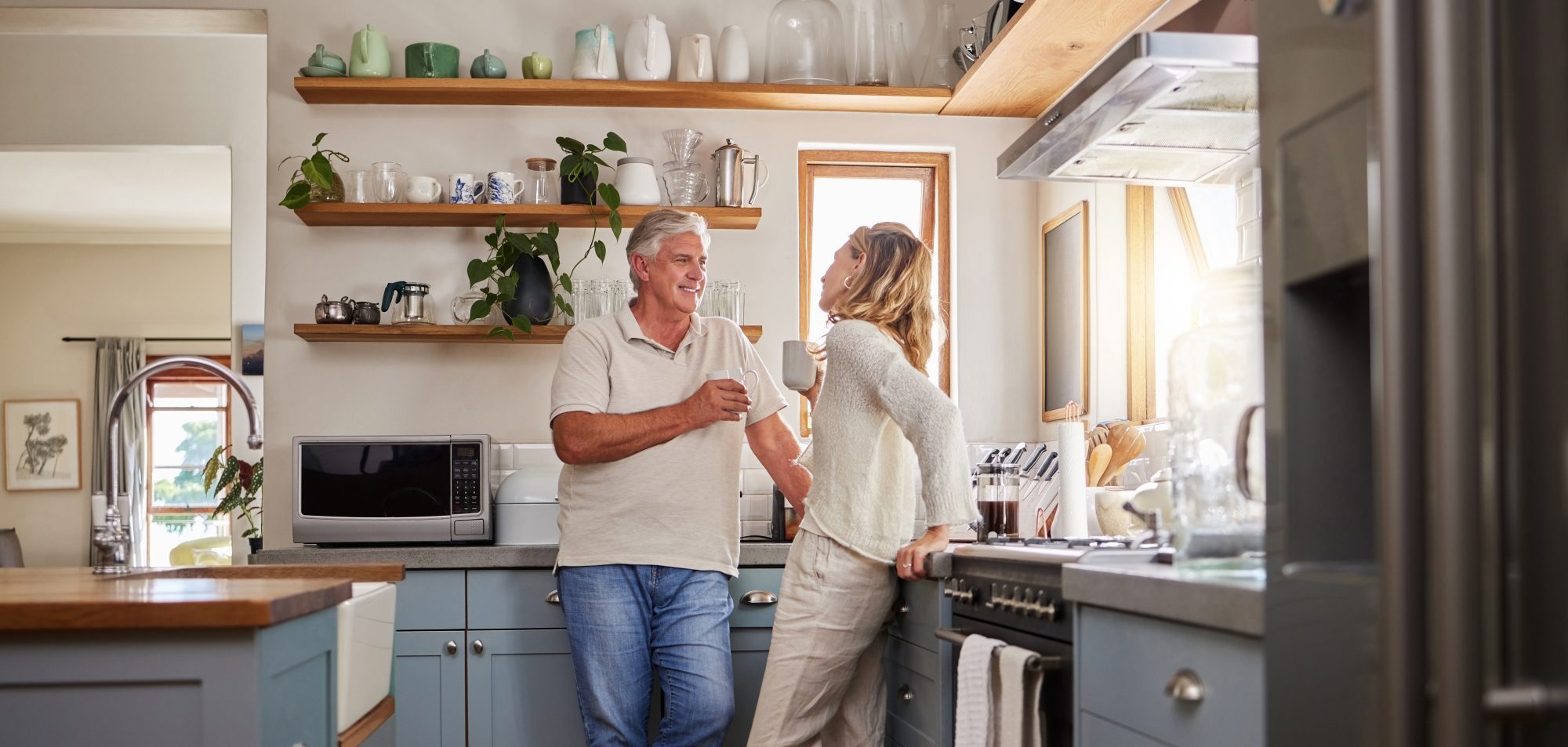 An older man and a woman stand in a modern kitchen with blue cabinets, talking and smiling while holding mugs, perhaps discussing advice from LaGrange elder law attorneys.