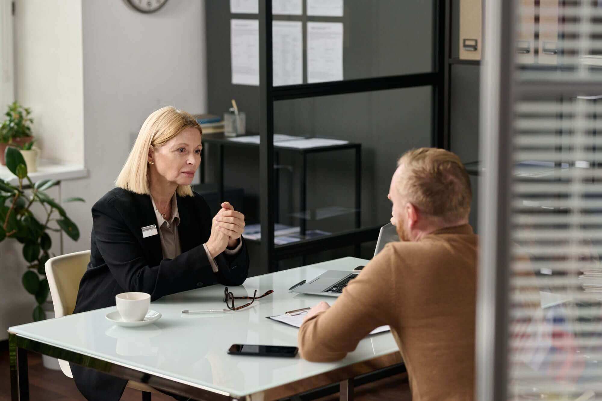Two people sit across a desk in an office setting, discussing LaGrange Estate Planning. A laptop, notebook, and cup are on the table. The room's glass walls and clock create a professional ambiance fitting for such important discussions.
