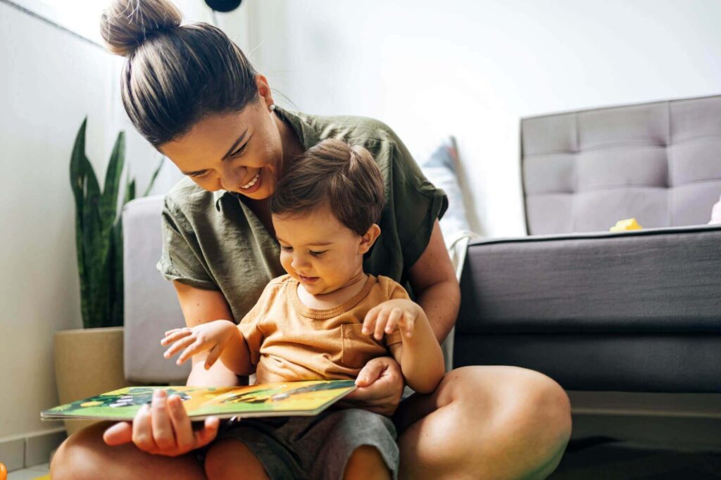 A woman and a child sit on the floor, reading a colorful book together. The child points at the book, and both are smiling. A couch and plants are in the background, evoking a warm family scene reminiscent of cherished moments in places like Western Springs Estate homes.