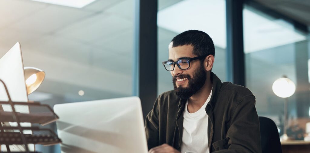 A man with glasses and a beard works at a laptop in a modern office, smiling as he types, with desk organizers and a lamp nearby.