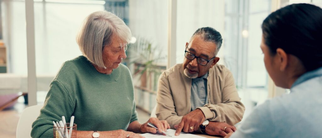Two older adults sit at a table reviewing and discussing documents, with another person partially visible in the foreground.