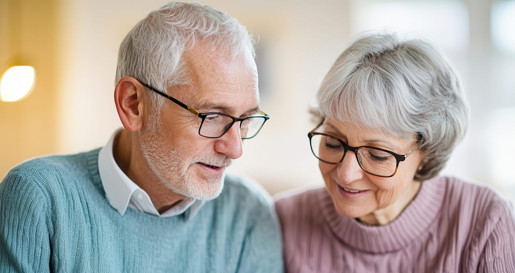 An elderly man and woman with glasses sit together, reviewing and discussing a document on a clipboard indoors.