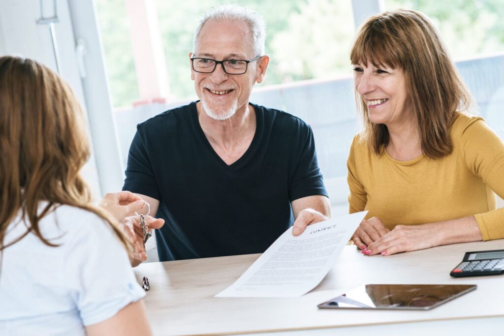 An elderly couple sits across from a woman at a table, discussing Hinsdale Wills. The man holds keys, while the woman holds a contract. A tablet and calculator are strategically placed on the table, aiding their important conversation about future planning.