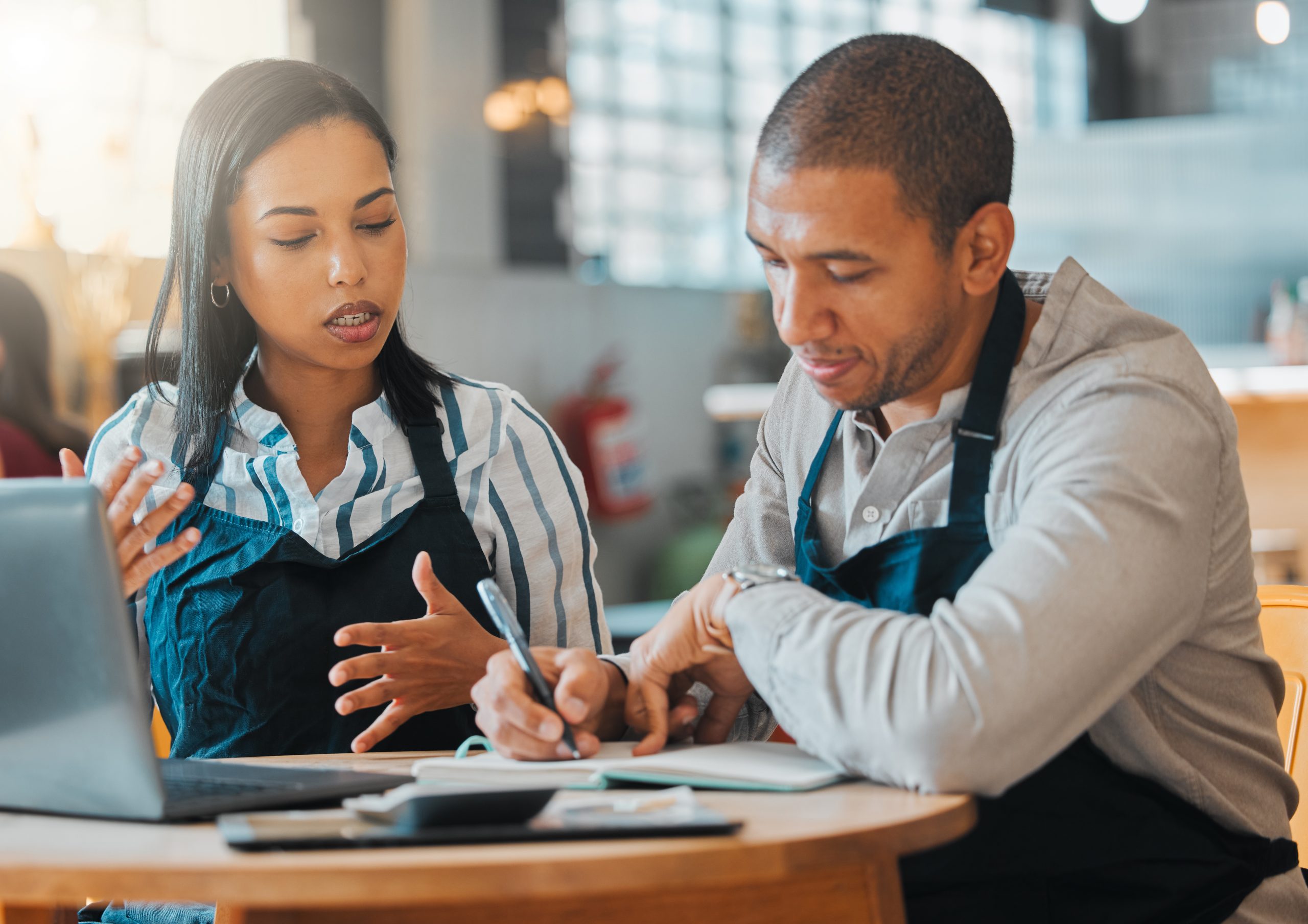 Two people wearing aprons sit at a table. One gestures while speaking, the other takes notes on Western Springs Estate Administration. A laptop is open in front of them, efficiently capturing their discussion.