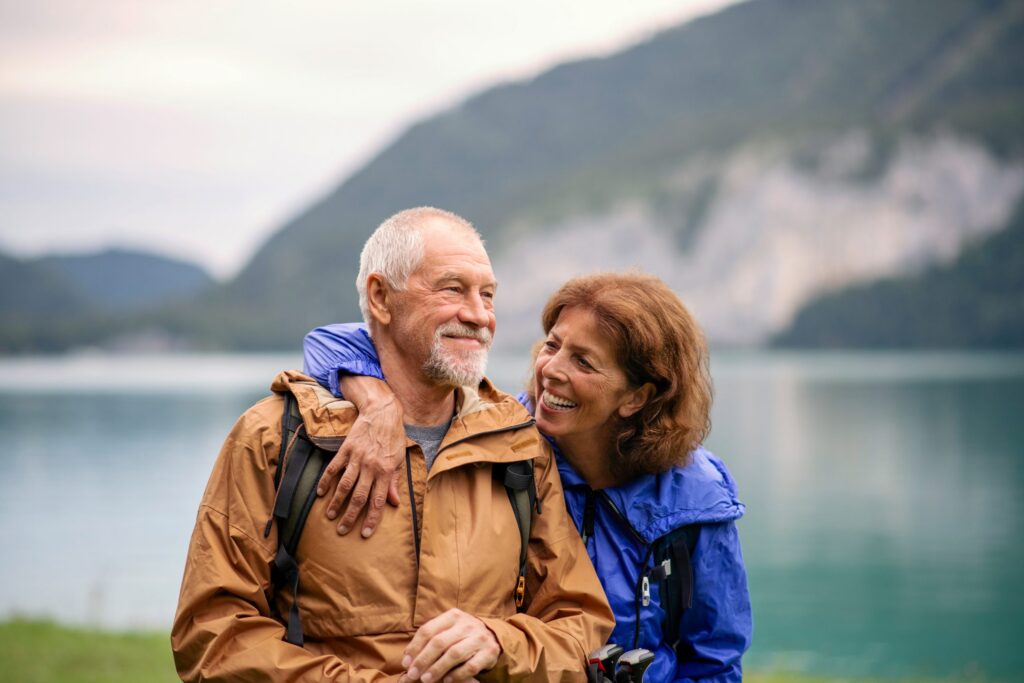 Elderly couple smiling outdoors by a lake, with mountains in the background, a serene scene reminiscent of peaceful days spent discussing LaGrange Park Trusts. The man wears a brown jacket, and the woman in blue embraces him warmly.
