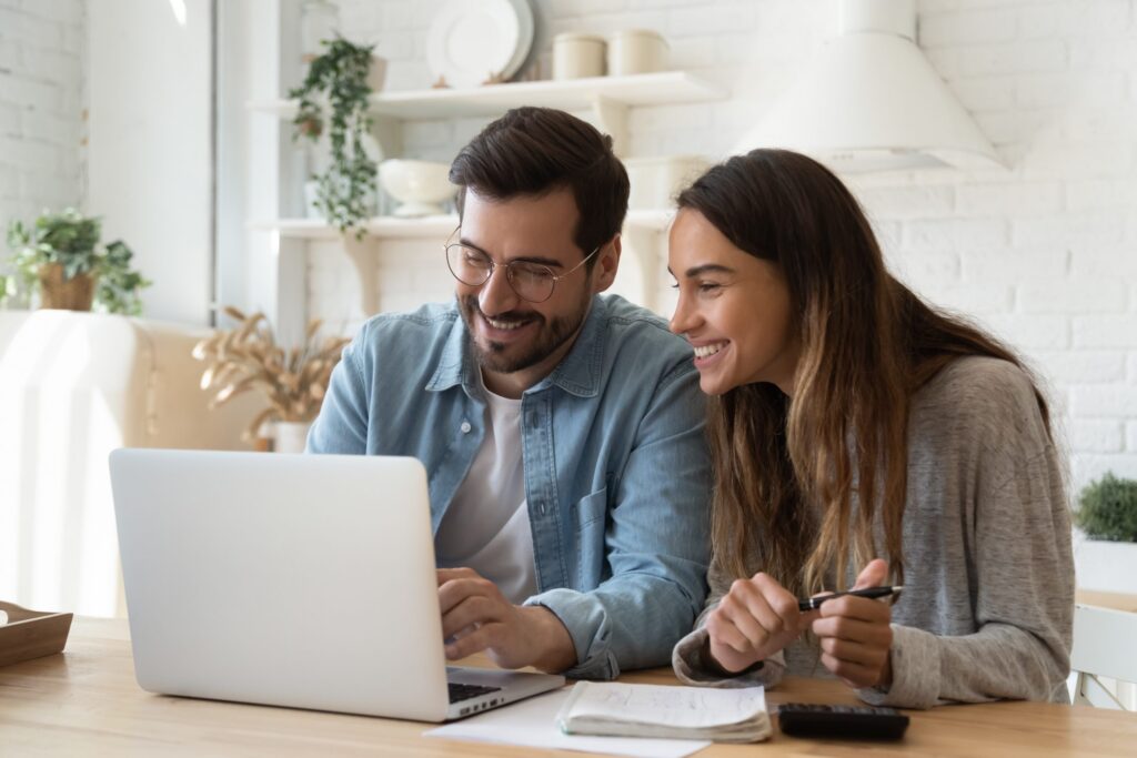 A man and woman sit together at a table, smiling as they look at a laptop. Nearby are notebooks and a pen, hinting at discussions on Hinsdale Wills in the brightly lit room.