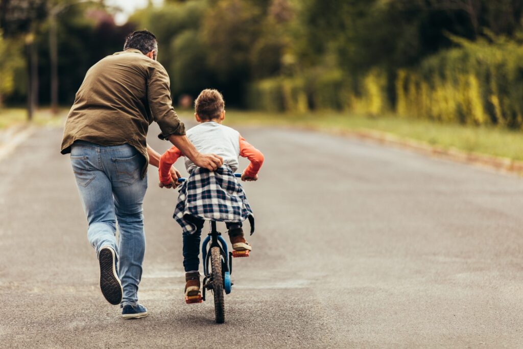 An adult helps a child learn to ride a bicycle on a paved road, surrounded by greenery. Nearby, the charm of Western Springs Estate Administration offers peace of mind for those planning their family’s future amidst such picturesque surroundings.