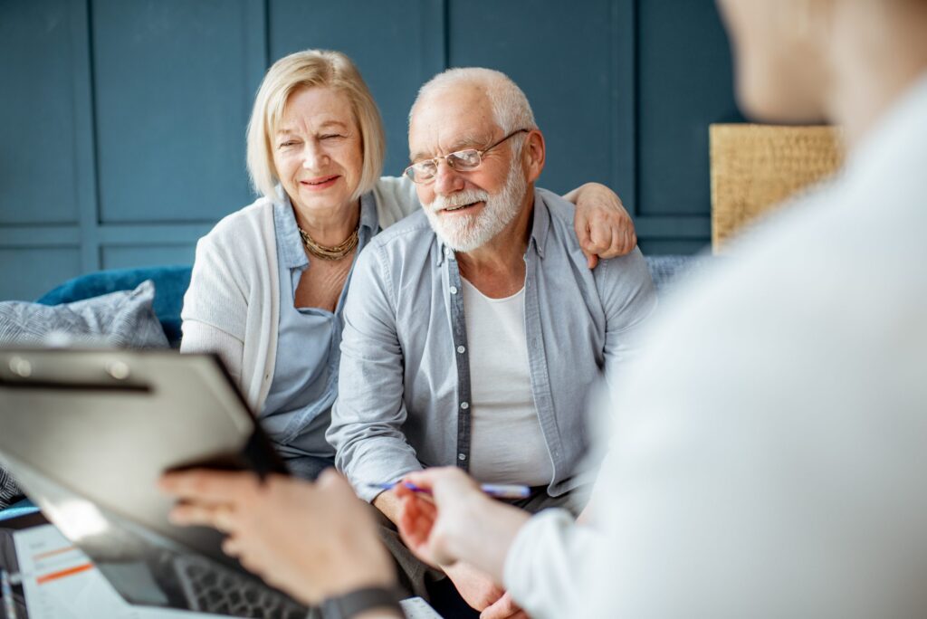 An elderly couple sits together on a couch, attentively looking at a person holding a clipboard with a pen in an office or consultation setting, discussing their plans with Brookfield Elder Law experts.