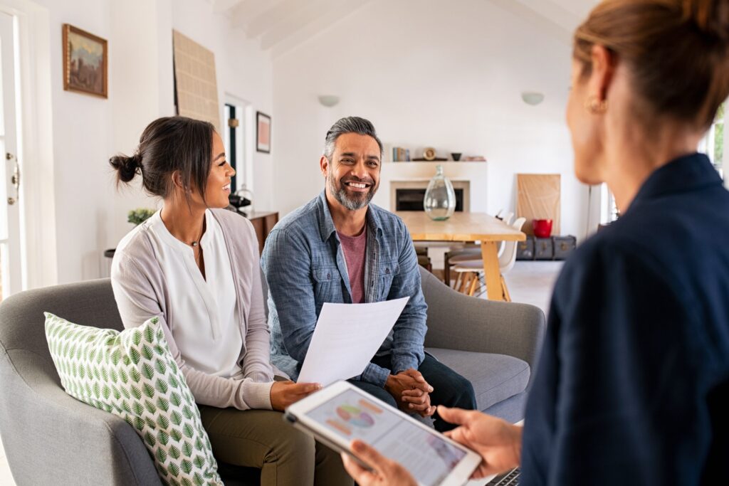 A couple sits on a couch with paperwork, smiling at a person holding a tablet. In their bright, modern living room, they're discussing the benefits of Hinsdale Wills for secure future planning.