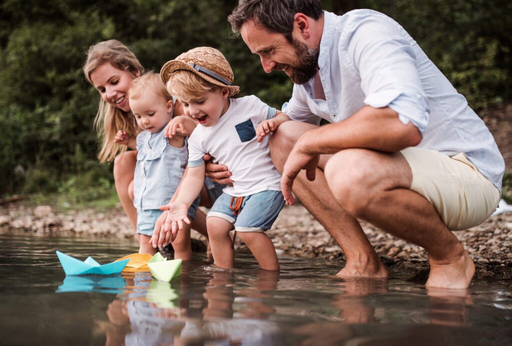 In a serene, wooded area, a family with two joyful children enjoys the simple pleasure of sailing paper boats in shallow water. This delightful scene mirrors the peacefulness often found in Western Springs estate administration proceedings.