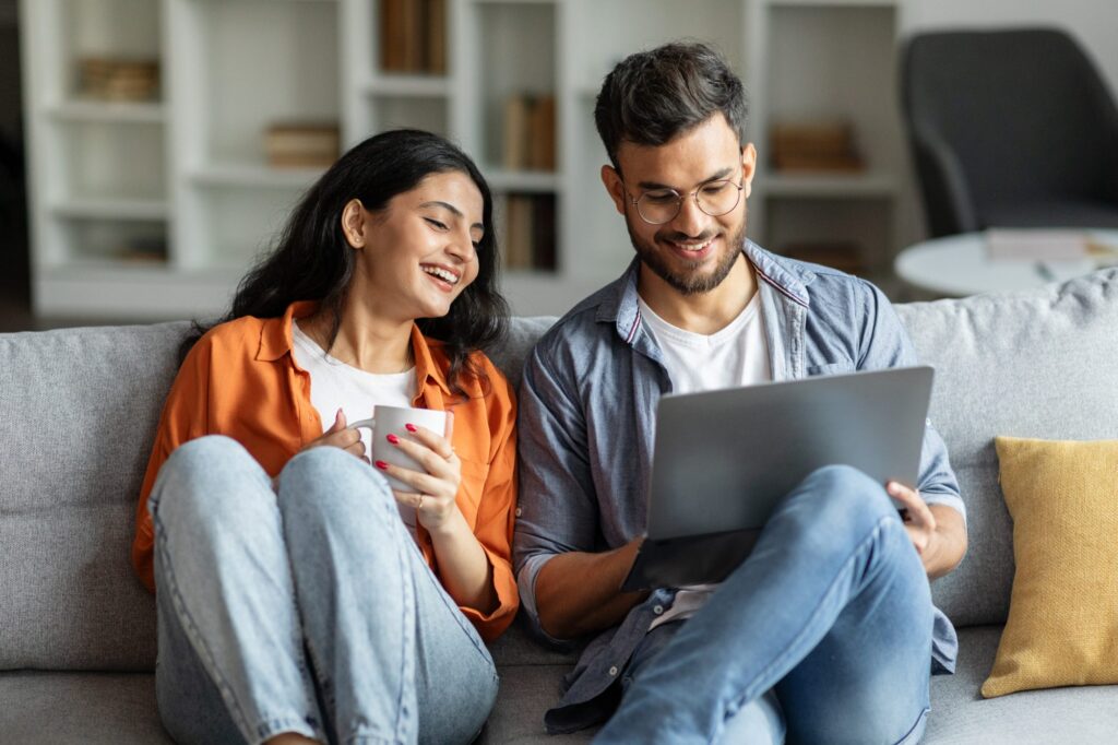 Two people sit on a couch. One holds a laptop, possibly researching Hinsdale Wills, while the other, holding a mug, listens intently. Both appear engaged and relaxed in their conversation.