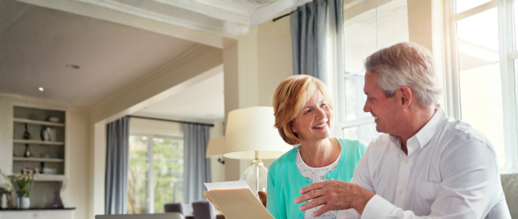 An older couple sits on a couch in a bright living room, holding documents and having a conversation, with a laptop on the table in front of them.
