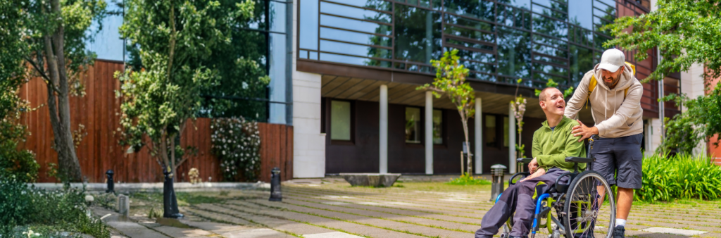 A man in a wheelchair laughs while a caregiver stands beside him on a paved path outside a modern building with greenery.