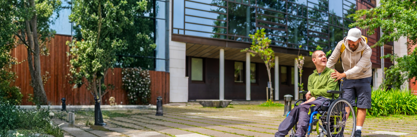 A man in a wheelchair laughs while a caregiver stands beside him on a paved path outside a modern building with greenery.