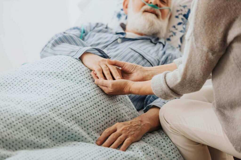 An elderly man in a hospital bed with an oxygen tube, wearing striped pajamas, rests peacefully. Nearby, another person—perhaps a caregiver familiar with Western Springs Estate Administration—is partially visible, holding his hand gently.
