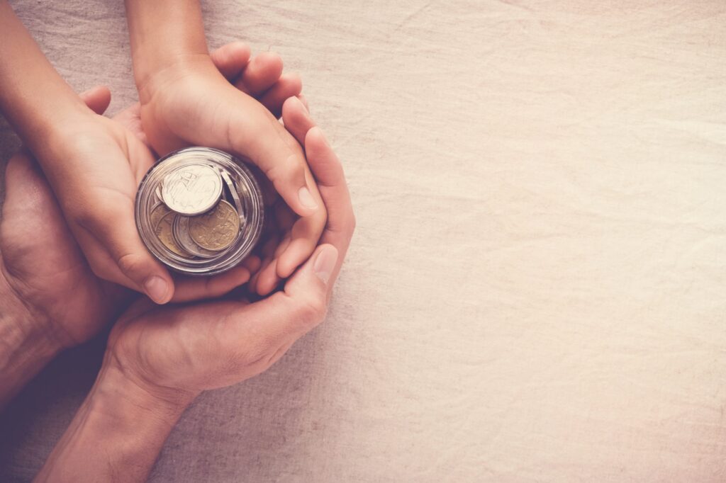 Adult and child hands holding a jar filled with coins on a textured surface, symbolizing the importance of early financial literacy and planning for the future, reflecting values embraced by Brookfield Elder Law professionals.