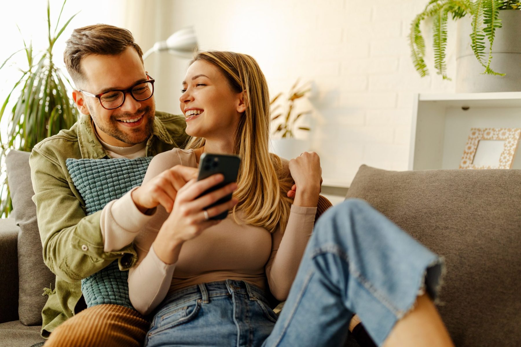 A couple sits on a couch, smiling as they look at a smartphone, possibly browsing LaGrange Estate Planning options. Plants and a lamp create a cozy atmosphere in the background.